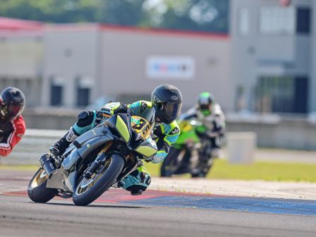 Three motorcyclists in racing gear take a sharp turn on a racetrack, with a building in the background.