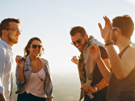 Four people are outdoors, laughing and dancing. They appear to be having a good time together. The background shows a bright sky.