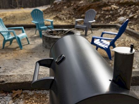 The image shows a black smoker grill in the foreground, with a fire pit surrounded by colorful Adirondack chairs in the background.