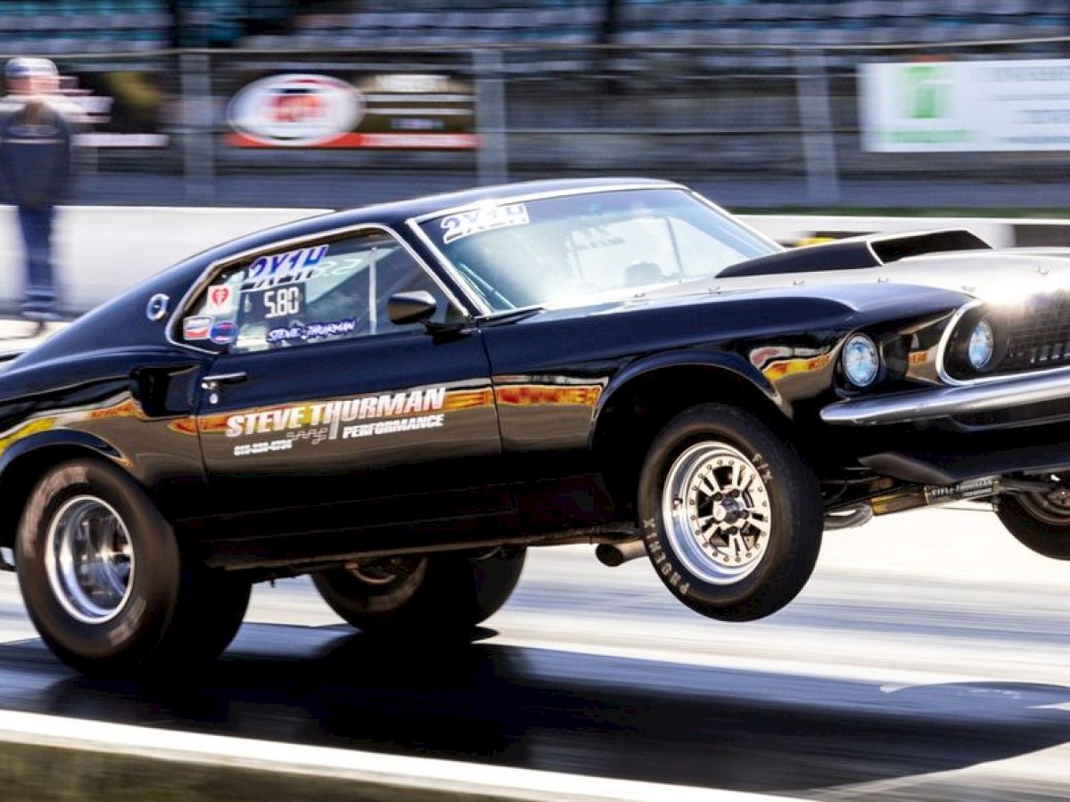 A black muscle car launching with its front wheels off the ground at a drag strip, with the name "Steve Thurman" and other decals on the sides.