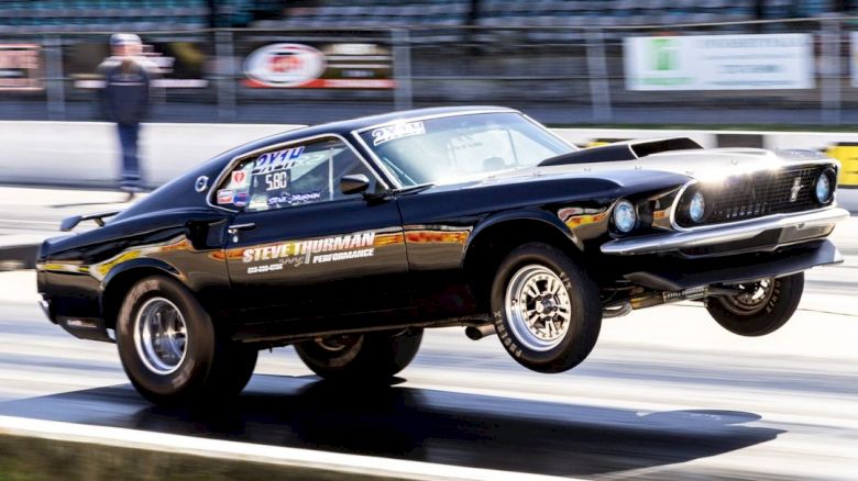 A black muscle car launching with its front wheels off the ground at a drag strip, with the name 