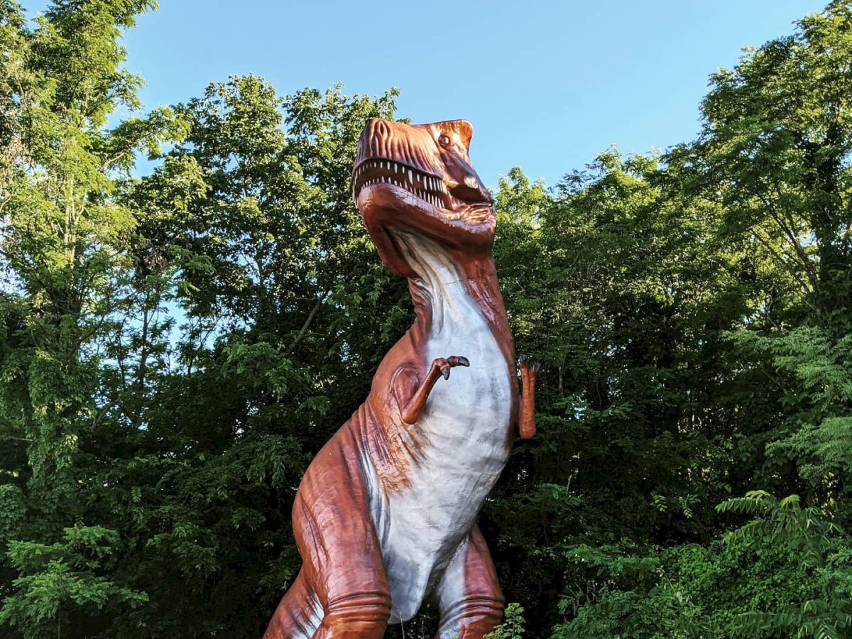 A large T-Rex statue is seen amongst lush green trees and vegetation, standing on its hind legs with an open mouth under a clear blue sky.