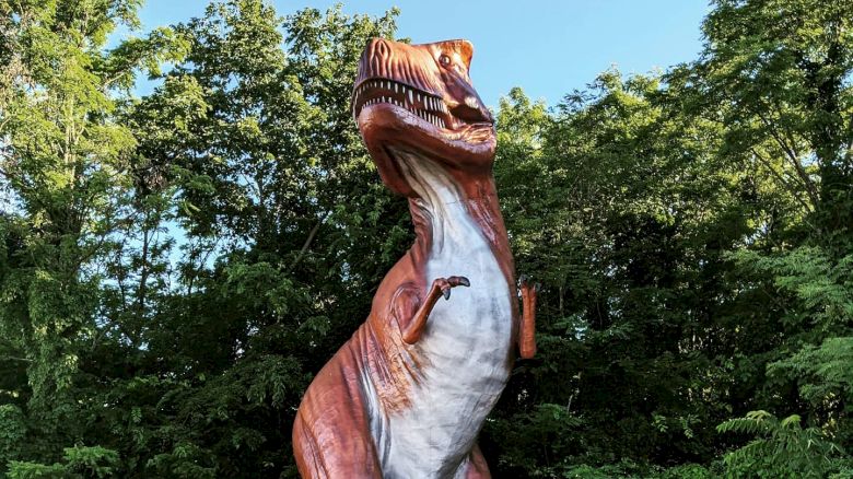 A large T-Rex statue is seen amongst lush green trees and vegetation, standing on its hind legs with an open mouth under a clear blue sky.