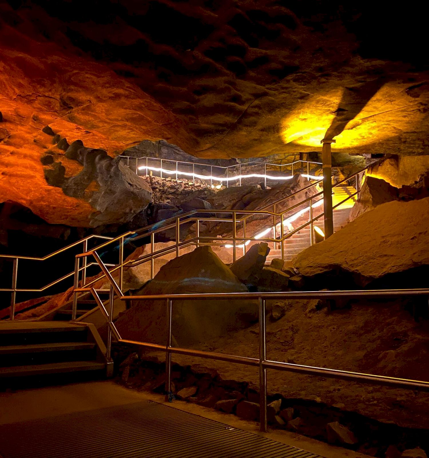 An illuminated cave interior with a winding staircase and railings leading upwards, surrounded by rock formations and warm lighting.