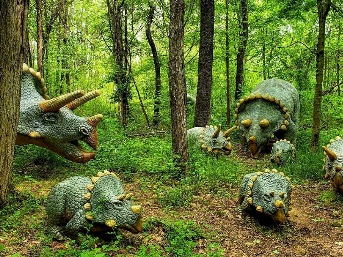 The image shows dinosaur statues in a forest setting, featuring several triceratops figures including adults and young ones amidst trees and greenery.