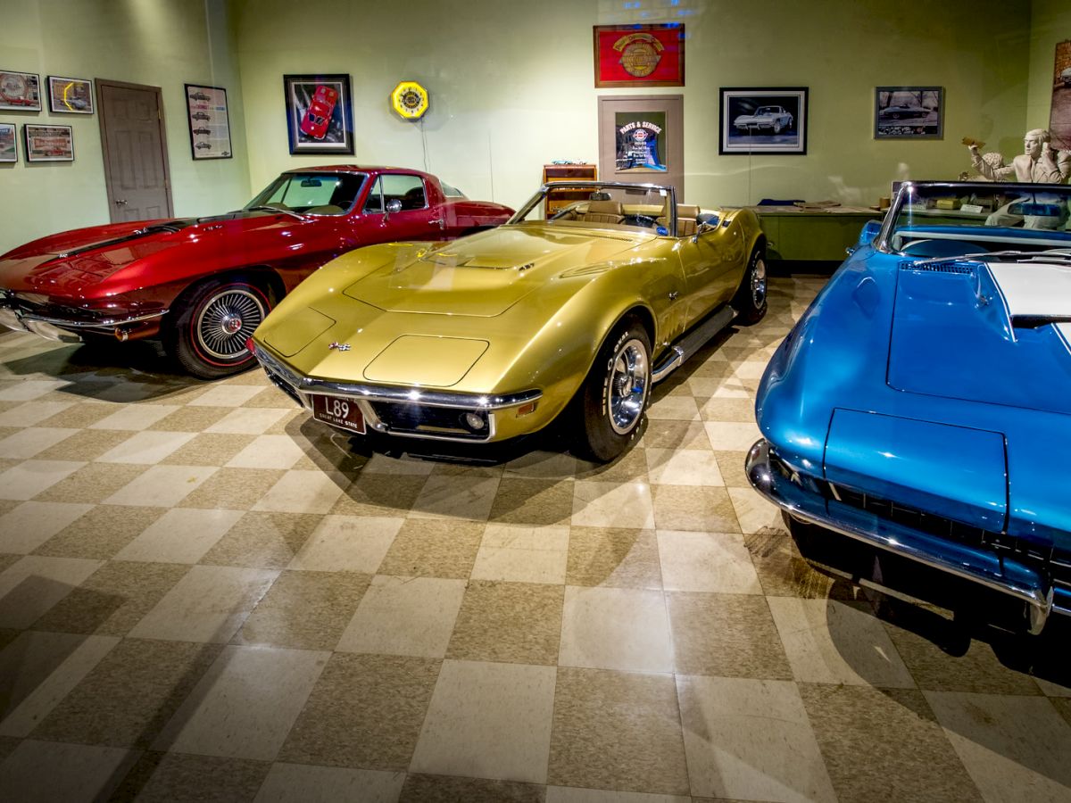 The image shows three classic cars in a showroom: a red car on the left, a yellow car in the center, and a blue car on the right.