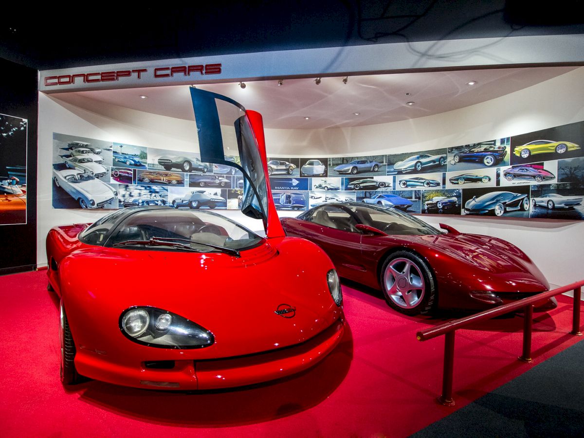 The image shows a display of two red concept cars in an exhibition with pictures of other car designs mounted on the wall behind.