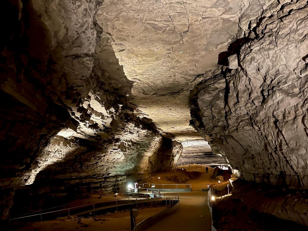 A well-lit cave passage with a winding walkway and rugged rock formations on the ceiling and walls, illuminated by strategically placed lights.