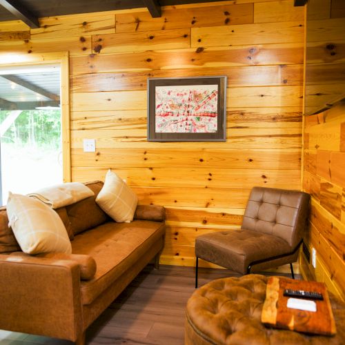 A cozy living space features a brown couch with pillows, a chair, an ottoman, framed artwork, wood-paneled walls, and a sliding glass door.