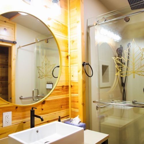 A modern bathroom with a round mirror, wooden walls, a white sink, and a glass shower door featuring a maple leaf design.