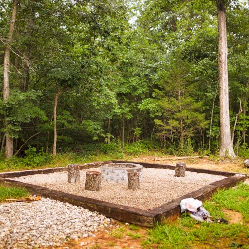 An outdoor fire pit with tree stump seats, surrounded by a wooded area, setup on a gravel platform with trees in the background, ending the sentence.