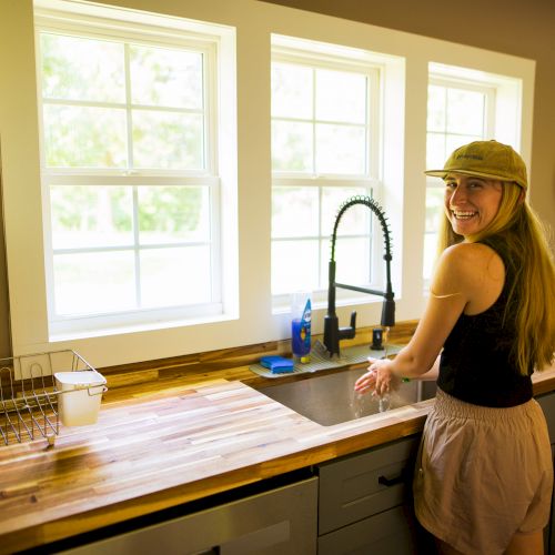 A person wearing a hat smiles while washing hands in a kitchen with a wooden countertop, next to a window with a view of greenery outside.