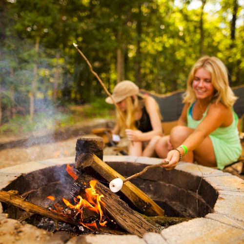 Two people are roasting marshmallows over a campfire within a wooded area, sitting on camping chairs. One of them is smiling.