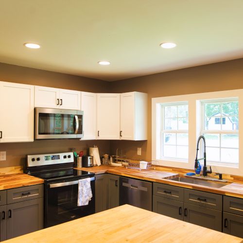 A modern kitchen with a wooden island, white upper cabinets, gray lower cabinets, stainless steel appliances, and a window above the sink.