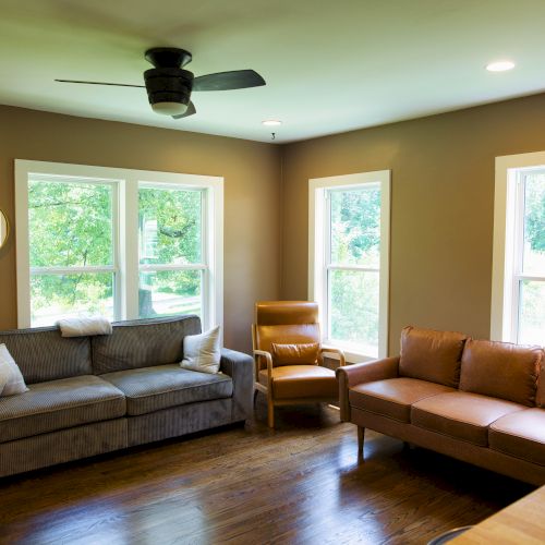 A cozy living room with two sofas, an armchair, large windows, a ceiling fan, and wooden flooring. Natural light fills the space.