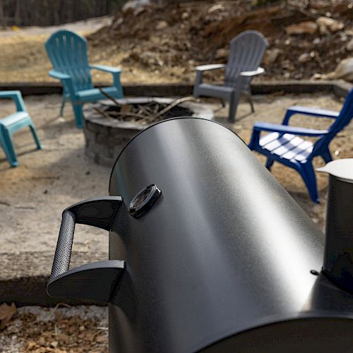 A black smoker grill in the foreground with a fire pit and several colorful Adirondack chairs arranged around it in the background.