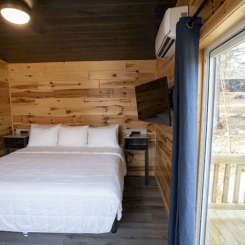 A cozy wooden cabin bedroom with a neatly made bed, wall-mounted TV, air conditioner, and large window opening to a deck with a forest view.