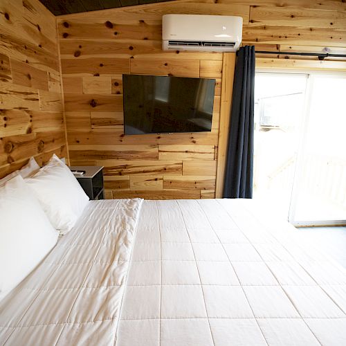 A cozy wooden room with a neatly made bed, wall-mounted TV, and an air conditioning unit, featuring curtains and a sliding door leading outside.