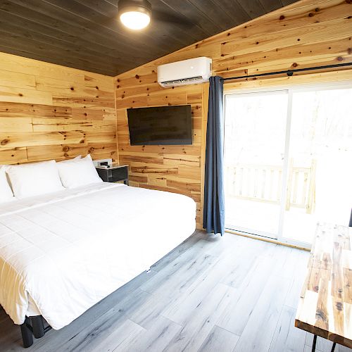 A cozy bedroom with wooden walls, a king-sized bed, TV, air conditioner, wooden table, and sliding glass doors with dark curtains opening to the outside.