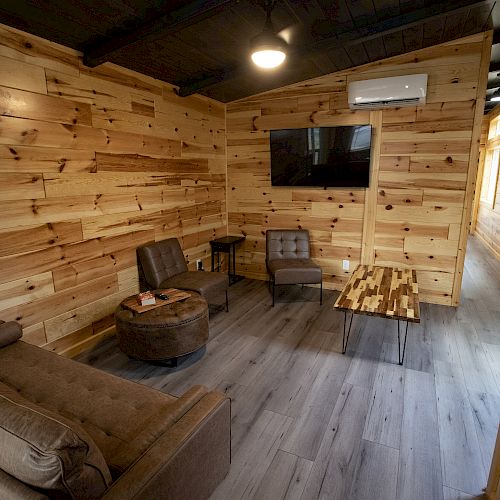 The image shows a cozy room with wooden walls, a gray floor, a couch, chairs, a TV on the wall, a coffee table, and a small table in the corner.