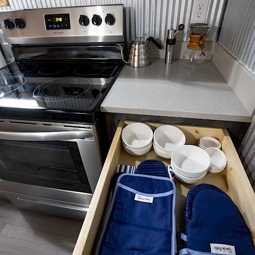 The image shows a kitchen with an open drawer containing blue oven mitts and white measuring cups, next to a stove and countertop.