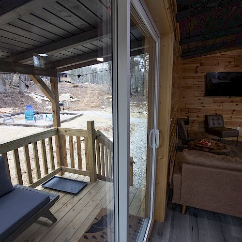 The image shows a cozy cabin interior with a sofa, TV, and a small porch with chairs, set against a wooded outdoor backdrop.