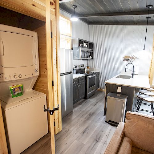 The image shows a small, modern kitchen and living area with a stacked washer and dryer, stainless steel appliances, and wooden accents throughout the space.