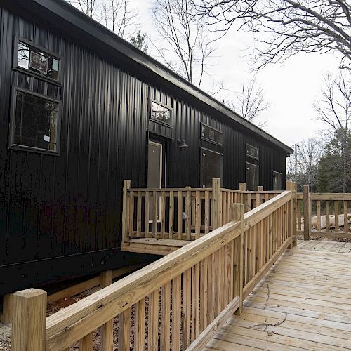 A long black metal container building with multiple windows, situated in a wooded area. The foreground features a wooden ramp leading to the entrance.