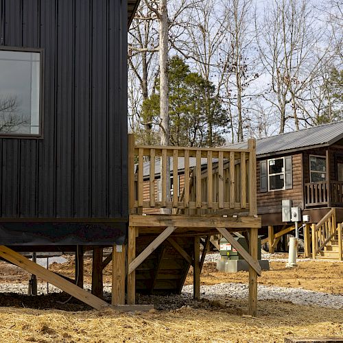 The image shows two elevated cabins in a wooded area with leafless trees. The foreground cabin is black, while the background cabin is brown.