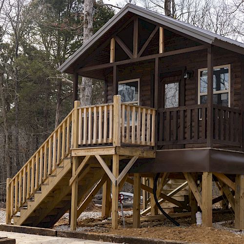 The image shows a small wooden cabin elevated on stilts, with a staircase leading up to a porch, set in a forested area during winter.