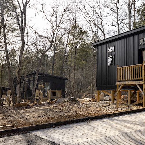 The image shows modern black cabins with wooden steps situated in a wooded area during daytime.