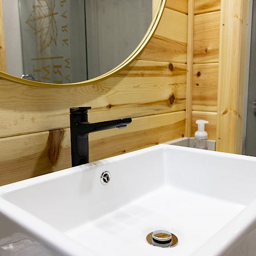 A bathroom with a modern white sink, black faucet, wooden wall, round mirror, soap dispenser, and a glass shower door is pictured.