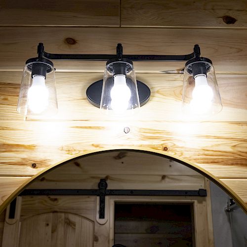 A wooden wall with three light bulbs in glass fixtures above a round mirror, reflecting a part of a door with a sliding barn door mechanism.