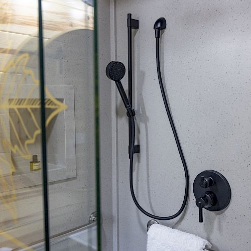 A modern bathroom shower with a matte black handheld showerhead, wall-mounted controls, a glass partition with a design, and a towel hanging.