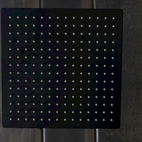 The image shows a black perforated square panel with evenly distributed holes, placed against a wooden background.