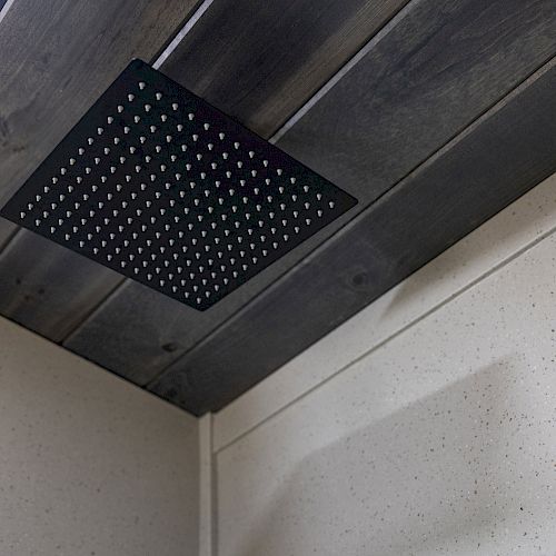 The image shows a black, square rainfall showerhead mounted on a dark wooden ceiling, with white tiled walls surrounding it.