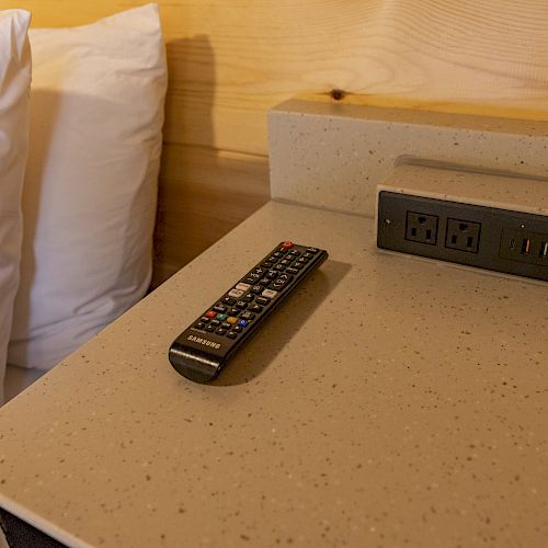 A bedside table with a remote control, electrical outlets, USB ports, and pillows in the background ends the sentence.