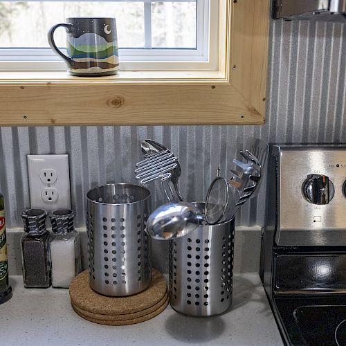 A kitchen counter with olive oil, salt and pepper, utensil holders, cork trivets, and a stainless steel stove. A mug is on the window sill.