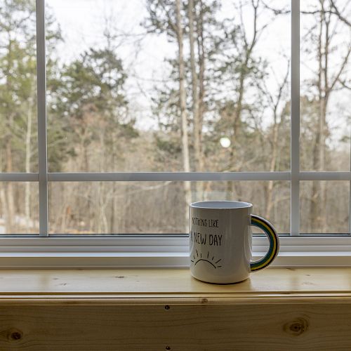 A white mug sits on a wooden windowsill in front of a window, with a view of trees and a cloudy sky in the background, creating a cozy scene.