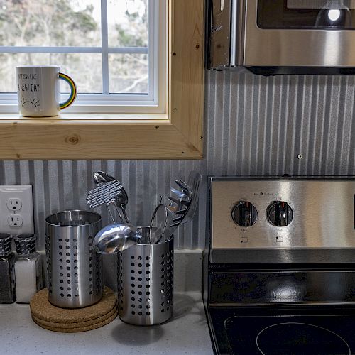 A kitchen scene with utensils, a mug on the window ledge, a bottle of olive oil, and part of a stove with a corrugated metal backsplash.