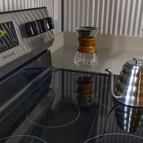 The image shows an electric stovetop with a stainless steel kettle and a pour-over coffee maker on the counter with a metallic wall background.