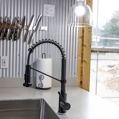 A modern kitchen sink with a black faucet, knife rack, paper towel holder, and a pendant light, next to a window overlooking an outdoor scene.