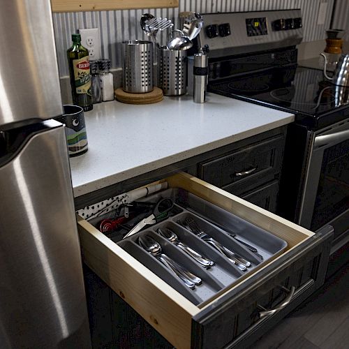 The image shows a modern kitchen with a stainless steel fridge, stove, utensils on the counter, and an open drawer containing silverware.