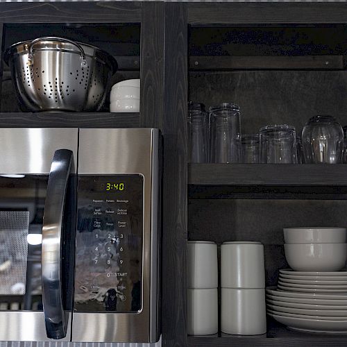 A kitchen with a microwave, colander, and shelves holding glasses, plates, bowls, and mugs.