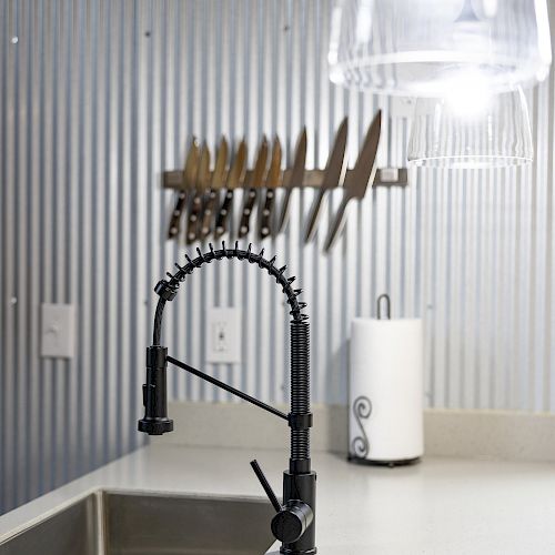 A kitchen scene with a black faucet, stainless steel sink, corrugated metal walls, knife holder, paper towel roll, and a hanging glass light fixture.