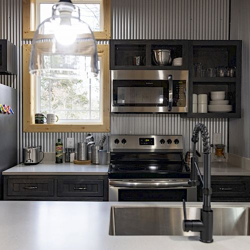 A modern kitchen with stainless steel appliances, dark cabinets, a white countertop, and a central sink under pendant lighting.