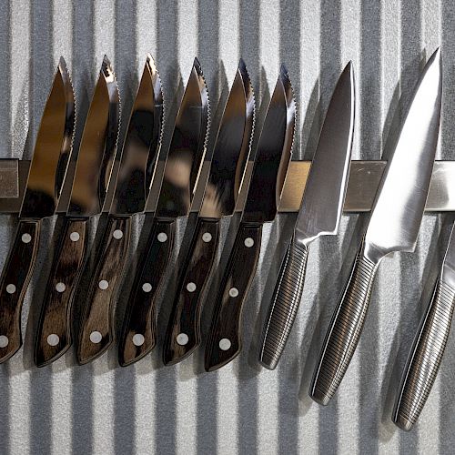 A collection of various kitchen knives is neatly arranged on a magnetic knife holder against a striped metal background.