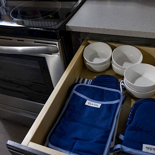 An open kitchen drawer with various white bowls and blue oven mitts next to a stainless steel oven and a countertop.