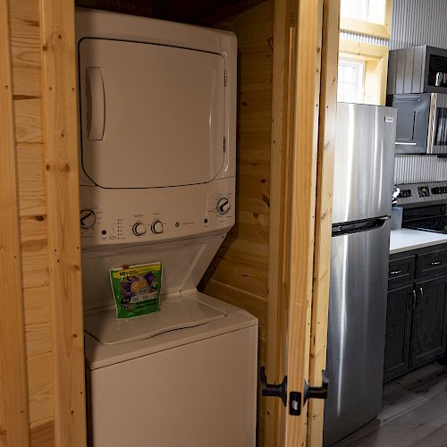 The image shows a compact laundry area with a stacked washer and dryer inside a wooden enclosure, adjacent to a kitchen with modern appliances.