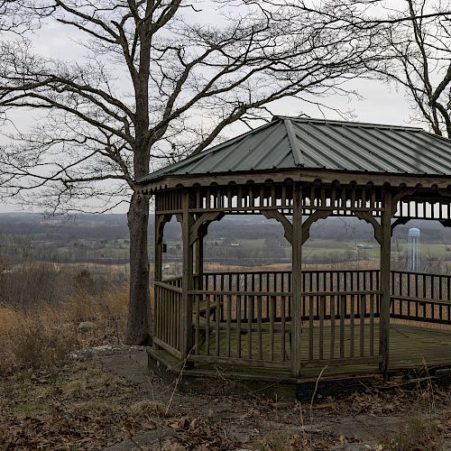 A wooden gazebo stands among bare trees with a rural landscape stretching into the distance; a calm and serene scene with autumn leaves.